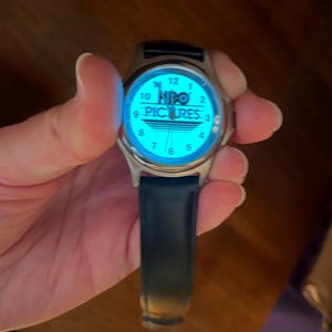 May include: A silver-toned wristwatch with a blue illuminated face. The watch face displays the text "HBO PICTURES" and standard hour markers. The watch has a dark blue leather band.
