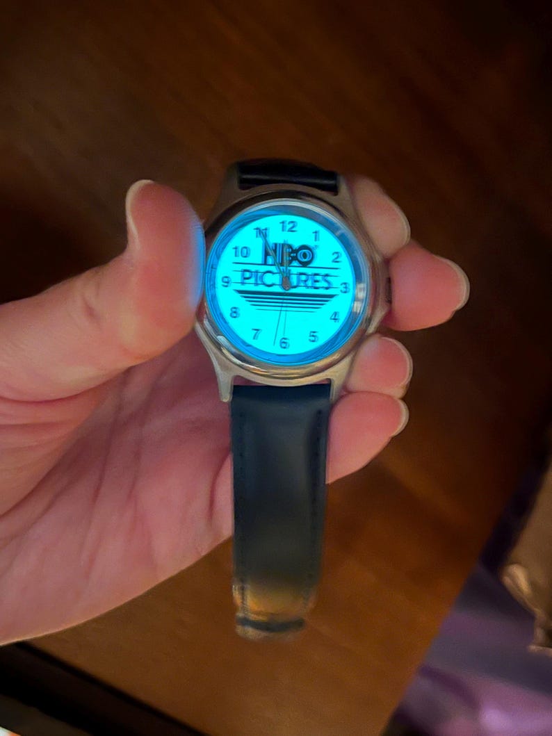 May include: A silver-toned wristwatch with a blue illuminated face. The watch face displays the text "HBO PICTURES" and standard hour markers. The watch has a dark blue leather band.
