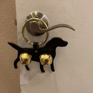 May include: A black leather dog-shaped door knocker with two gold bells attached. The knocker is hanging on a silver door handle.