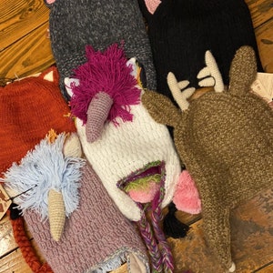 May include: A collection of five knit hats featuring animal designs including a dark gray cat, a black cat, a white unicorn with a magenta mane, a rust-colored fox, and a brown deer with antlers. Each hat includes earflaps and braided tassels.