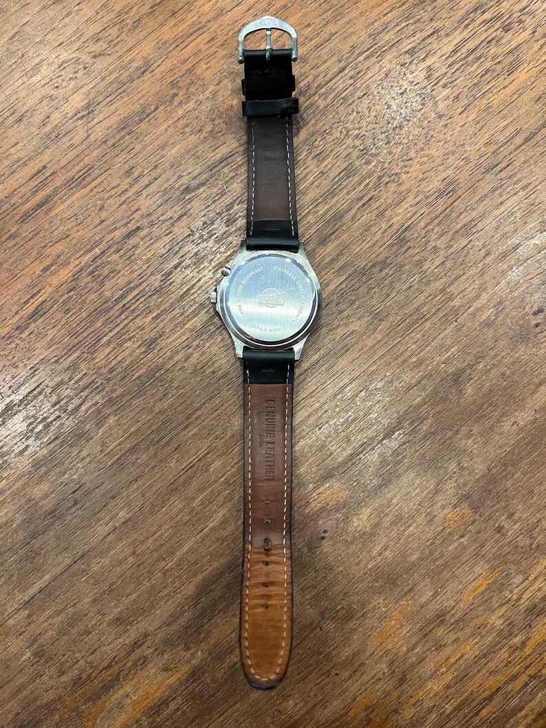 May include: A silver-toned wristwatch with a black leather band. The watch face is not visible, but the back of the watch is visible, with text and a logo. The band is marked "Genuine Leather".