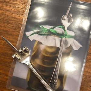 May include: Two silver serving forks rest on a photograph of a jar of pickles. One fork has a hand-shaped design, the other has a decorative handle. The photograph is on a wooden surface.