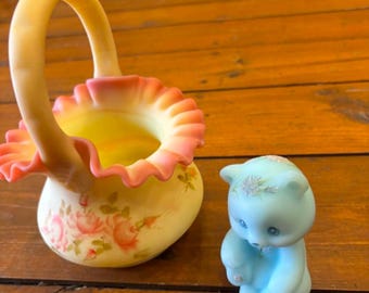 May include: A yellow glass basket with a ruffled pink rim and painted pink rose floral details, accompanied by a small light blue bear figurine featuring a floral accent on its head and a small white flower detail on its chest.