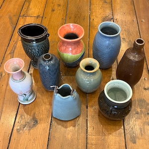 May include: A collection of handmade ceramic vases in various shapes, sizes, and colors. The vases include shades of blue, brown, pink, and green. Some have textured surfaces, while others are smooth. The collection is arranged on a wooden surface.