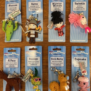 May include: Ten colorful string doll keychains with different characters and names. Each keychain is packaged on a card with a description of the character. The characters include a cactus, a warrior woman, a pineapple, a squirrel, a cupcake, a alpaca, a gunna, a twinkle, and a blossom.
