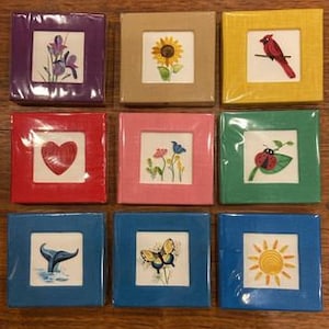 May include: Nine small square frames with colorful backgrounds and embroidered images. The images include a heart, a butterfly, a sunflower, a cardinal, a ladybug, a whale tail, a sun, and two floral designs.