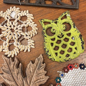 May include: A collection of decorative items including a cast iron trivet, a beaded coaster, a wooden leaf, and a distressed green owl-shaped trivet. The items are arranged on a wooden surface.