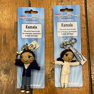May include: Two string doll keychains featuring Kamala Harris. The dolls are dressed in blue suits and white suits. The packaging includes the text "The String Doll Gang" and "Kamala" with a quote: "You never have to ask for someone's permission to lead. Just lead."