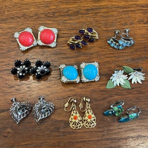 May include: A collection of vintage earrings in various styles. Includes red, blue, and black gemstone studs, gold filigree drop earrings, leaf-shaped silver earrings, and floral clip-ons. The earrings are displayed on a wooden surface.