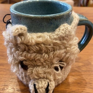 May include: A blue ceramic mug featuring a beige crocheted cozy shaped like an alpaca face. The cozy has textured yarn, two small ears, dark brown eyes, and a stitched black nose and mouth. The mug has a glossy finish and a curved handle.