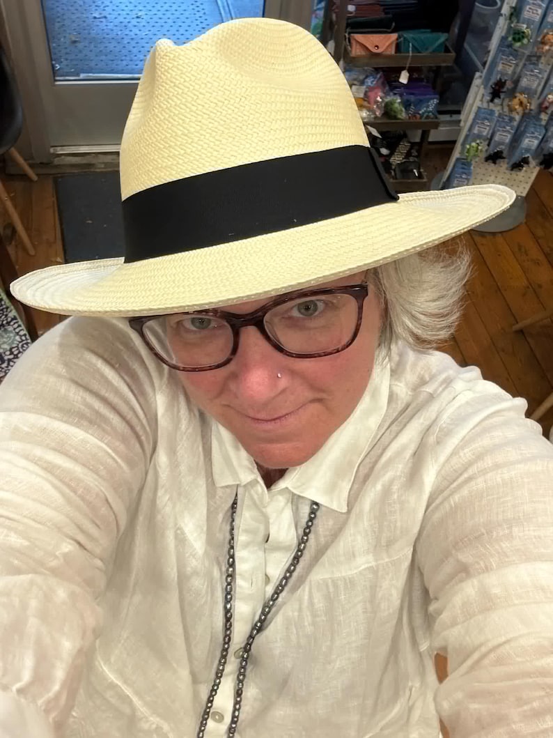 Puede incluir: Una persona que lleva una camisa blanca y un sombrero de paja con una banda negra. El sombrero tiene un ala ancha y est&aacute; hecho de un material tejido.