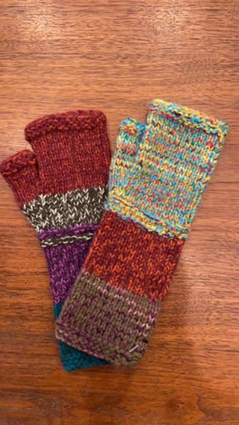 Op de afbeelding: Een paar kleurrijke vingerloze handschoenen met een gestreept patroon. De handschoenen zijn gemaakt van een zachte, gebreide stof en hebben een verscheidenheid aan kleuren, waaronder rood, oranje, geel, groen, blauw en paars.
