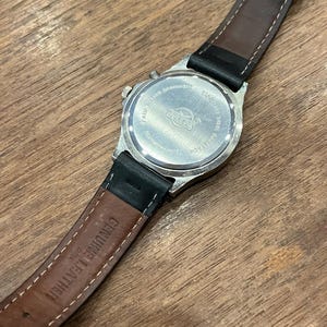 May include: A silver-toned watch with a black leather band. The watch face is not visible, but the back shows the brand name "SWISS" and other details. The band is brown on the inside and has "GENUINE LEATHER" printed on it.