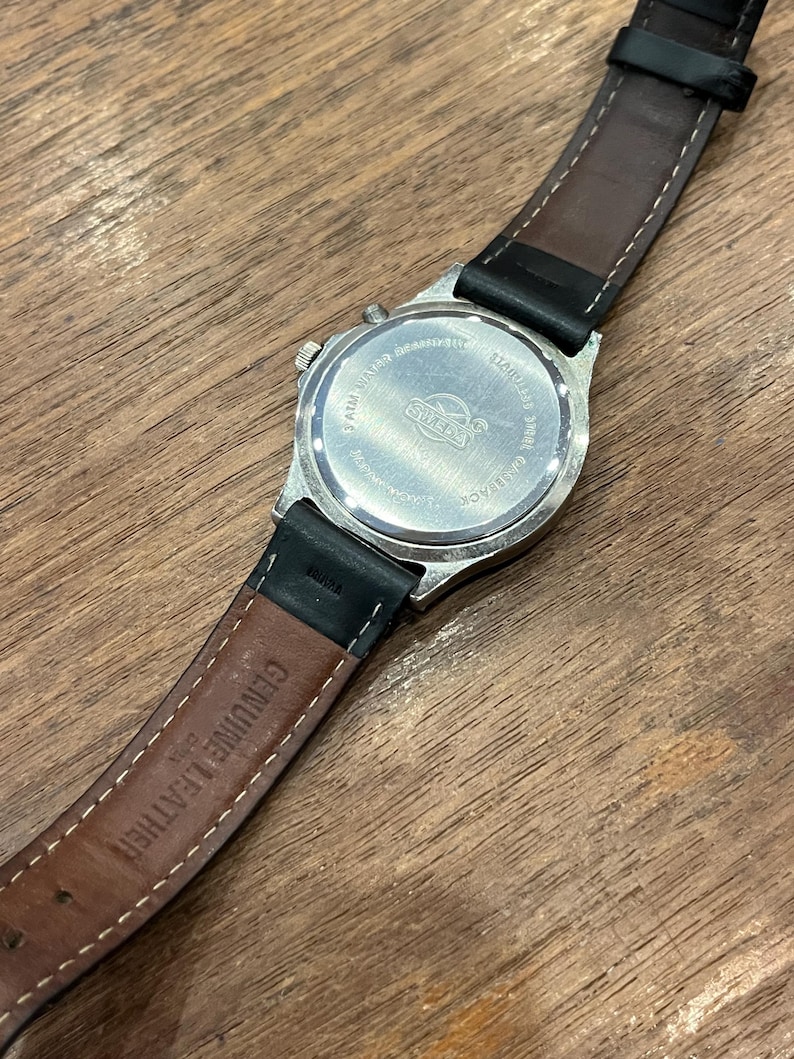 May include: A silver-toned watch with a black leather band. The watch face is not visible, but the back shows the brand name "SWISS" and other details. The band is brown on the inside and has "GENUINE LEATHER" printed on it.