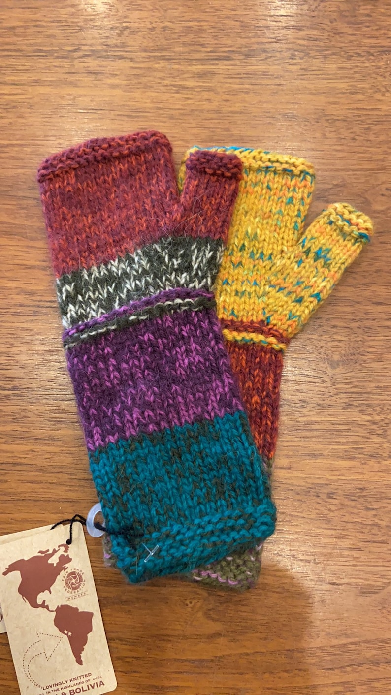 Op de afbeelding: Een paar kleurrijke vingerloze handschoenen met een gestreept patroon in tinten rood, paars, turquoise en geel. De handschoenen zijn gemaakt van gebreide wol en hebben een label met de tekst "Met liefde gebreid in de hooglanden van Bolivia".