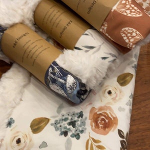 May include: Three fabric swaddles with a white faux fur trim. The swaddles are rolled up and tied with a brown paper band. The bands have the text "Kark Designs" printed on them. The swaddles have different floral patterns.