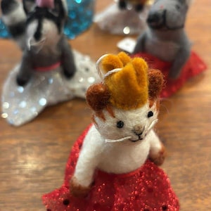 May include: Handmade felt animal figurines, including a lion with a gold crown and red skirt, and other animals with various accessories. The figurines are white, brown, gray, and red, with glitter accents. The background is a wooden surface.