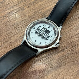 May include: A wristwatch with a white face featuring the text "PIC RES" and black numerals. The watch has a silver-colored case and a black leather band. The watch is displayed on a wooden surface.
