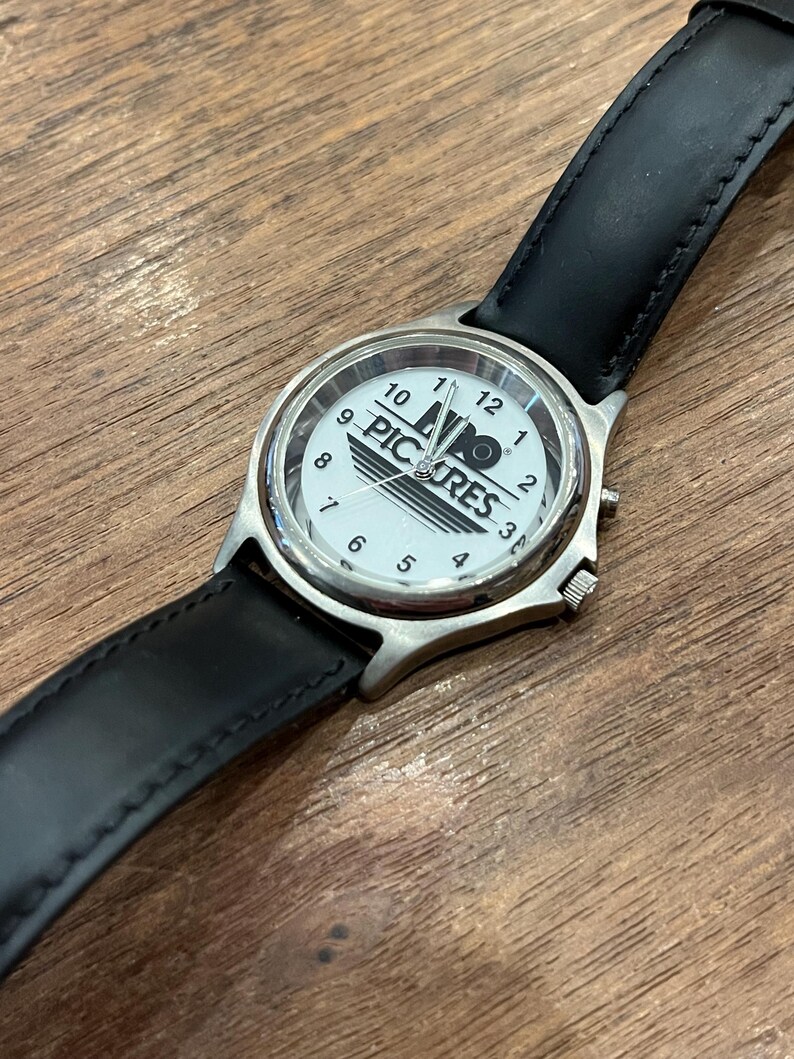 May include: A wristwatch with a white face featuring the text "PIC RES" and black numerals. The watch has a silver-colored case and a black leather band. The watch is displayed on a wooden surface.