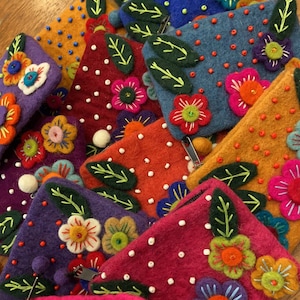May include: A stack of colorful felt coin purses with floral embroidery and beaded accents. Each purse features a different floral design with green leaves and white beads.