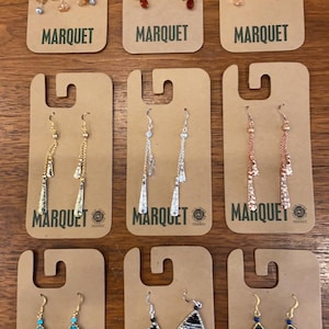 May include: Nine pairs of earrings displayed on brown card stock with the word "MARQUET" printed on each card. The earrings are various styles and colors, including silver, gold, and beaded.