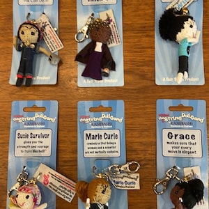 May include: Six string doll keychains featuring historical figures. Each doll is attached to a blue card with the doll's name and a quote. The dolls include Rosie the Riveter, Harriet Tubman, Willow, Susie Survivor, Marie Curie, and Grace.