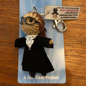 May include: A brown yarn doll with black glasses and a black robe, wearing a white lace collar. The doll is attached to a silver key chain with a silver clasp. The doll is on a blue and white background with the text "A Fair Trade Product" and "Women belong in all places where decisions are being made." The text "kamibashi.com" is also visible.
