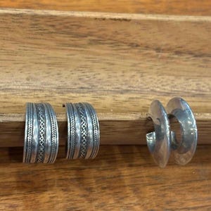 May include: Two pairs of silver hoop earrings. One pair features an etched design with intricate patterns, while the other pair has a smooth, polished finish. The earrings are displayed on a wooden surface.