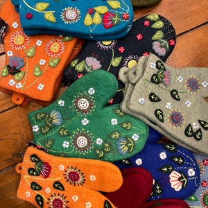 May include: A collection of colorful felt oven mitts with embroidered floral designs. The mitts are in various colors, including orange, green, blue, red, and gray. Each mitt features a unique floral pattern with leaves, flowers, and buttons.