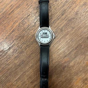 May include: A wristwatch with a white face and black leather band. The watch face features black numerals and the text "NIO PICTURES". The watch has a silver-colored metal casing and a buckle closure.