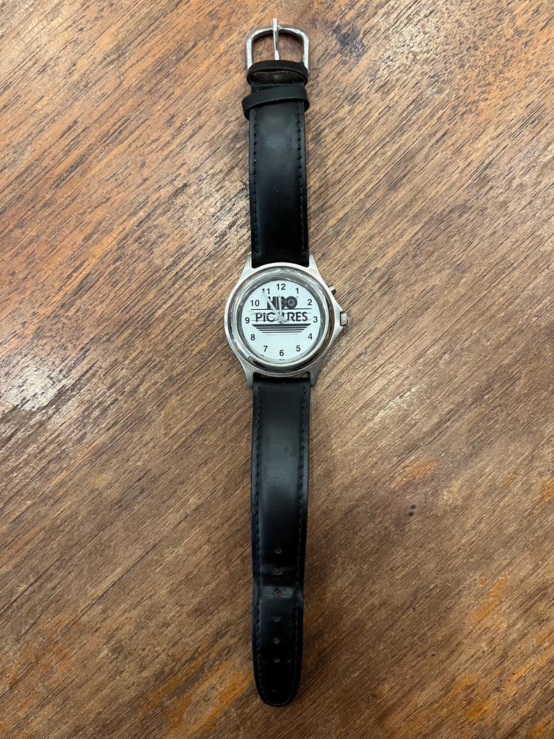 May include: A wristwatch with a white face and black leather band. The watch face features black numerals and the text "NIO PICTURES". The watch has a silver-colored metal casing and a buckle closure.
