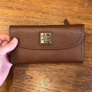 May include: A brown leather wallet with a flap closure. The wallet features a textured surface and a gold-colored metal plaque with the brand name "Dooney & Bourke" embossed on it. The wallet is approximately 7 inches long.