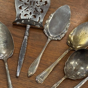 May include: A collection of five silver-toned metal utensils including a decorative pierced server, a pointed cake server with an ornate border, and three various spoons with different handle patterns and levels of tarnish on the bowls.