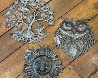May include: Three circular metal wall art pieces with a weathered silver finish. One depicts a tree with two birds, another shows a detailed owl face with textured feathers, and the third features a sun with a face surrounded by radiating rays.