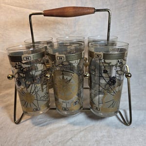 May include: A vintage set of six clear drinking glasses with a gold and black map design, held in a gold-colored metal carrier with a wooden handle. Each glass features the letter "N".