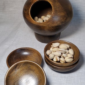 May include: A wooden nut bowl set with a large spherical bowl on a pedestal, a small bowl, and two small bowls stacked. The large bowl is filled with pistachios. The smaller bowls also contain pistachios.