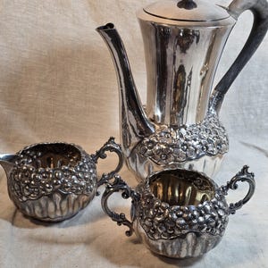 May include: A silver-plated tea set consisting of a tall coffee pot with a dark handle and lid finial, a creamer, and a sugar bowl. Each piece features a prominent embossed floral pattern around the base and a fluted design on the lower body.