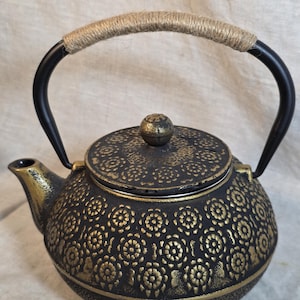 Könnte beinhalten: Eine schwarze gusseiserne Teekanne mit einem sich wiederholenden goldenen Blumenmuster auf dem Körper und dem Deckel. Die Teekanne hat einen geschwungenen schwarzen Metallgriff, der mit natürlichem Jutegarn umwickelt ist, und einen kleinen, runden, strukturierten Knopf in der Mitte des Deckels.