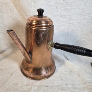 May include: A copper coffee pot with a cylindrical body, a long angled spout, and a black wooden handle. The pot features a matching copper lid topped with a small black knob. The metal surface shows signs of wear and natural oxidation.