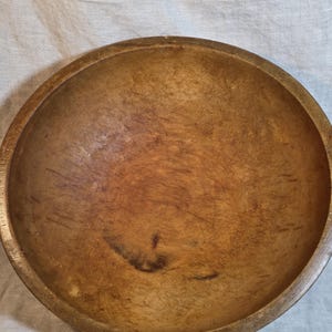 May include: A large, round wooden bowl with a natural light brown finish. The interior surface displays visible grain patterns, subtle variations in wood tone, and numerous small, shallow surface marks throughout the basin of the bowl.