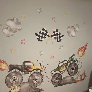 May include: Wall stickers depicting two monster trucks with flames and dirt effects. The design includes checkered flags, clouds, and star accents, creating a dynamic scene. The trucks are in shades of black, yellow, and green, suitable for a child's room.