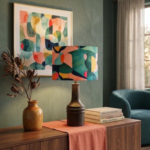 Peut inclure: Une lampe de table avec une base cylindrique en métal foncé et un abat-jour tambour présentant un motif abstrait de formes bleu sarcelle, corail, jaune et bleu marine. La lampe est posée près d'un vase en céramique beige avec des branches séchées.