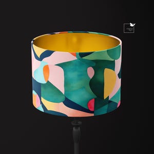 Puede incluir: Una pantalla de lámpara colorida con formas abstractas en tonos de rosa, verde, azul, amarillo y naranja. La pantalla tiene un interior dorado y está hecha de tela.