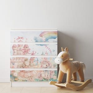 May include: A white dresser with four drawers featuring a whimsical watercolor design of a princess, a unicorn, a castle, and a rainbow. The dresser is decorated with pink, blue, and green colors. A rocking horse is in the foreground.