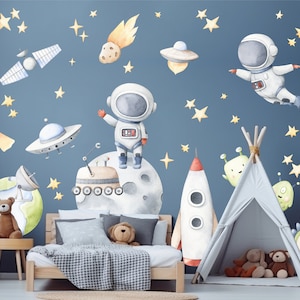 Può includere: Un set di adesivi murali a tema spaziale con illustrazioni ad acquerello di astronauti, un razzo, una luna, pianeti, un satellite, UFO e stelle gialle su sfondo blu, disposti sopra un letto con struttura in legno e cuscini.