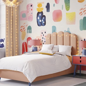 Op de afbeelding: Een lichtroze, beige en blauwe gestoffeerde bed met een gebogen hoofdeinde en een wit dekbed. Het hoofdeinde heeft een string van lichtsnoeren eroverheen gespannen. De muur achter het bed heeft een kleurrijke abstracte muurschildering met roze, blauwe, groene en gele vormen.