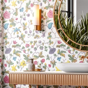 May include: A white bathroom with a floral wallpaper featuring a variety of flowers and leaves in shades of pink, yellow, blue, and green. A gold vanity with a white porcelain sink and a gold mirror with a round frame are visible. A gold wall sconce with a white light fixture is mounted on the wall.