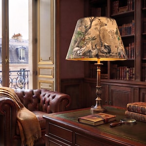 May include: A table lamp with a metallic base and a conical lampshade featuring a detailed print of cranes in a natural landscape. The lamp stands on a wooden desk alongside a leather-bound book and a magnifying glass in a library setting.