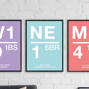 May include: Three framed posters with black borders. Each poster has a different background colour: lilac, pale blue, and salmon. White text indicates a location code and street name. The posters are displayed against a white brick wall.