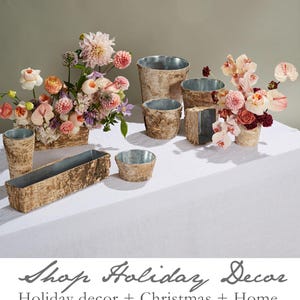 May include: A collection of birch bark planters in various sizes and shapes, perfect for adding a rustic touch to your home decor. The planters are filled with colorful flowers, including pink roses, white orchids, and purple dahlias. The text "Shop Holiday Decor" is written in cursive at the bottom of the image.